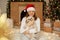 Adorable happy girl in red hat hugging with cute Pekingese dog on background of christmas tree with lights, fireplace and gift