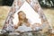 Adorable little blond girl wearing dress and hair feathers, sitting in a boho tent teepee.