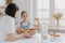 Adorable little child in apron shows whisk with white cream, wears apron, cooks together with mommy, pose in spacious kitchen