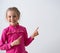 Adorable little girl in bright rose dress smiling and pointing aside with both her index fingers. Waist-up shot isolated