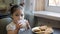 Adorable little girl is eating cheesecakes with her hands at the table in kitchen. Glass of yogurt on the table