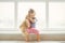 Adorable little girl hugging a teddy bear. Cute baby at home in white room is sitting near window.