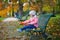 Adorable little girl sitting on the bench with lunchbox and having picnic on a fall day