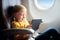 Adorable little girl traveling by an airplane. Child sitting by aircraft window and using a digital tablet