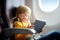 Adorable little girl traveling by an airplane. Child sitting by aircraft window and using a digital tablet during the flight.