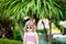 Adorable little girl at the tropical resort, standing by the palm three at the sunny summer day