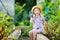 Adorable little girl wearing straw hat and childrens garden gloves playing with her toy garden tools in a greenhouse