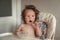 Adorable long haired toddler sitting in a baby high chair and eating porridge indoor.