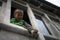 Adorable and playful young boy climb and sit at window ledge of home, looking down and leg stuck out.