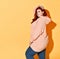 Adorable plus-size model posing in stylish everyday wear looking over shoulder and touching her red hair, isolated on orange