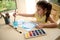 Adorable preschooler girl washes the paintbrush in a glass with water while painting picture with watercolor paints.