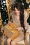 Adorable pretty teen girl with gift box over Christmas background. Happy funny brunette with long braid hairstyle sitting on mode