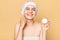 Adorable smiling woman applies moisturising mask, doing skin care procedures, puts nutritious cream, wearing shower cap and