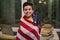 Adorable teenage schoolboy wrapped in American flag, stands by table with laptop and stacked workbooks, smiles at camera