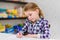 Adorable thoughtful little girl with blond hair sitting at table and drawing with purple pencil