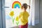 Adorable toddler girl attaching drawing of rainbow to window glass as sign of hope