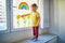 Adorable toddler girl attaching drawing of rainbow to window glass as sign of hope
