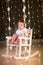 Adorable toddler girl in a white rocking chair in dark room with Christmas lights