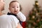 Adorable toddler sucking fingers standing by christmas tree on mother arms at home