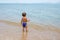 Adorable toddler toddler stands on the sandy beach and looks at the tropical sea.