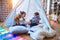 Adorable toddlers lying down over blanket inside tipi smiling and playing with doll at kindergarten