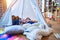 Adorable toddlers lying down over blanket inside tipi smiling and playing with doll at kindergarten