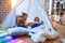 Adorable toddlers lying down over blanket inside tipi smiling and playing with doll at kindergarten