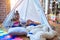 Adorable toddlers lying down over blanket inside tipi smiling and playing with doll at kindergarten