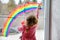 Adoralbe little toddler girl with rainbow painted with colorful window color during pandemic coronavirus quarantine