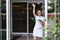 An adult beautiful woman in a white dress waves to someone, a neighbor or a guest client of a cafe, standing in the doorway. a
