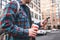 Adult casual man walking with cup og hot coffee on city street and texting on smartphone