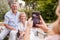 Adult Daughter Taking Picture Of Senior Parents Sitting In Garden On Mobile Phone