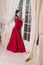 Adult elegant brunet 30 year old woman in long red dress stands by big window door. Vintage style classic beige interior