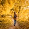 Adult girl walking away alone on path in autumn forest. Lonely young woman with backpack in beige autumn jacket