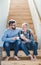 An adult hipster son and senior father sitting on stairs indoors at home, talking.