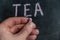 Adult male holding a piece of colored chalk in his hand. Handwritten word TEA on black chalkboard