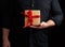 Adult man in a black shirt holds in his hand a square brown boxlet tied with a red ribbon
