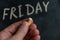 An adult man holds a piece of yellow chalk in his hand. Handwritten word FRIDAY on black chalkboard. Days of the week. Calendar