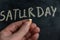 An adult man holds a piece of yellow chalk in his hand. Handwritten word SATURDAY on black chalkboard. Days of the week. Calendar