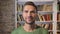 Adult man looking forward, smiling and blinking calmly. Bookshelves on the background