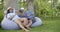 Adult pair of spouses are sitting in garden with palms on beanbag chair and drinking champagne