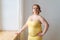 Adult plus size woman in tight-fitting yellow dress, makeup standing by window