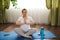 Adult pregnant woman practicing pregnany yoga at home, sitting on blue fitness mat, meditating in lotus position