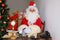 Adult santa claus in red suit sits at table, Christmas tree is beautifully decorated with balls, garlands, concept of christmas,