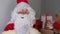 Adult santa claus in red suit sits at table, Christmas tree is beautifully decorated with balls, garlands, concept of christmas,