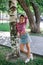 Adult shining blonde woman with face looking into camera standing in park leaning on birch wearing checkered shirt tied