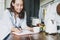 Adult smiling brunette woman in casual doing notes in daily book with cup of tea on kitchen