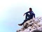 Adult tourist in black trousers, jacket and dark cap sit on cliff`s edge and looking into mist