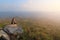 Adult tourist in black trousers, jacket and dark cap sit on cliff\'s edge and looking misty hilly valley bellow