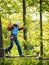 Adult woman in a helmet and with a safety system cautiously walking on suspension bridge on the background of trees